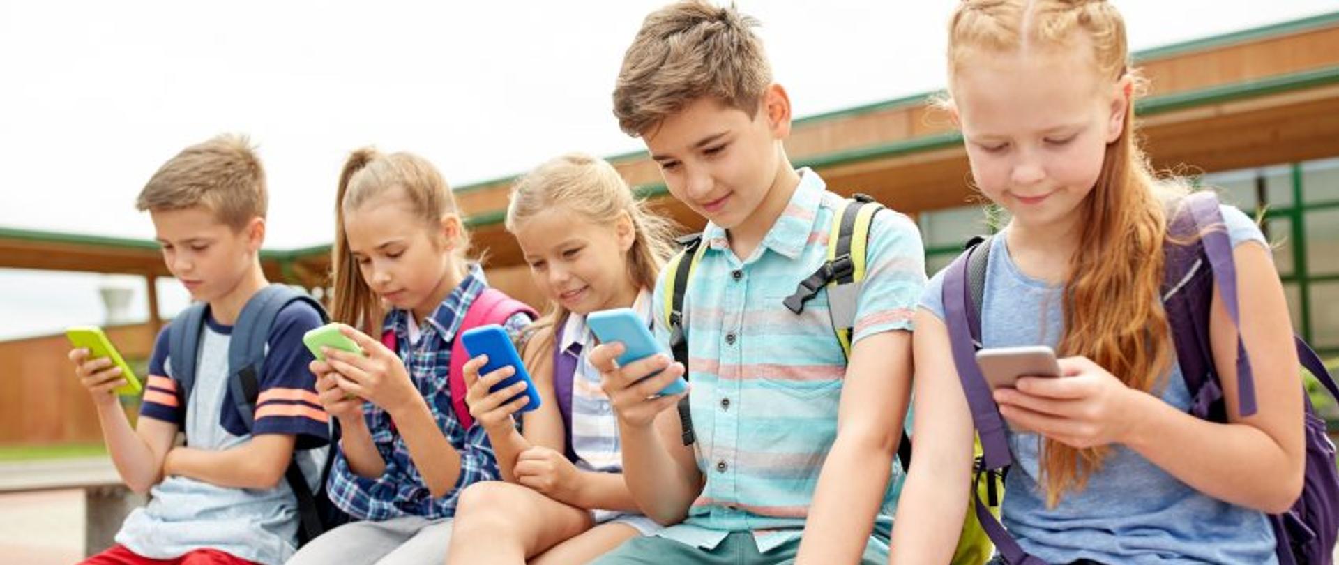 Dzieci siedzą na ławce. W ręku trzymają smartfony.