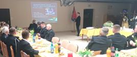 Strażacy siedzący przy stole podczas zebrania sprawozdawczego OSP. W tle widoczny strażak referujący informację o OSP w oparciu o prezentację multimedialną