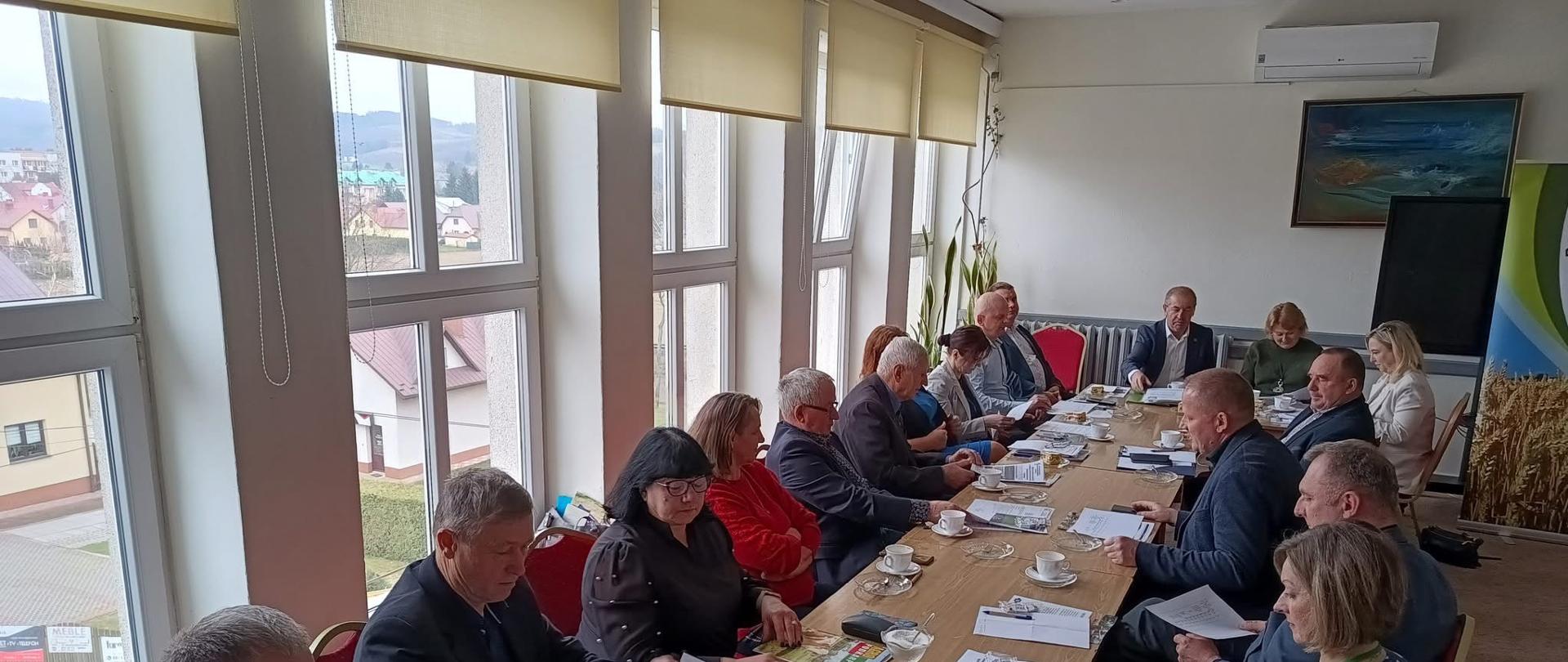 Kobiety i mężczyźni siedzą przy stole na którym leżą kartki papieru i filiżanki. Grupa ludzi znajduje się w jasnym pomieszczeniu w którym jest dużo okien.
