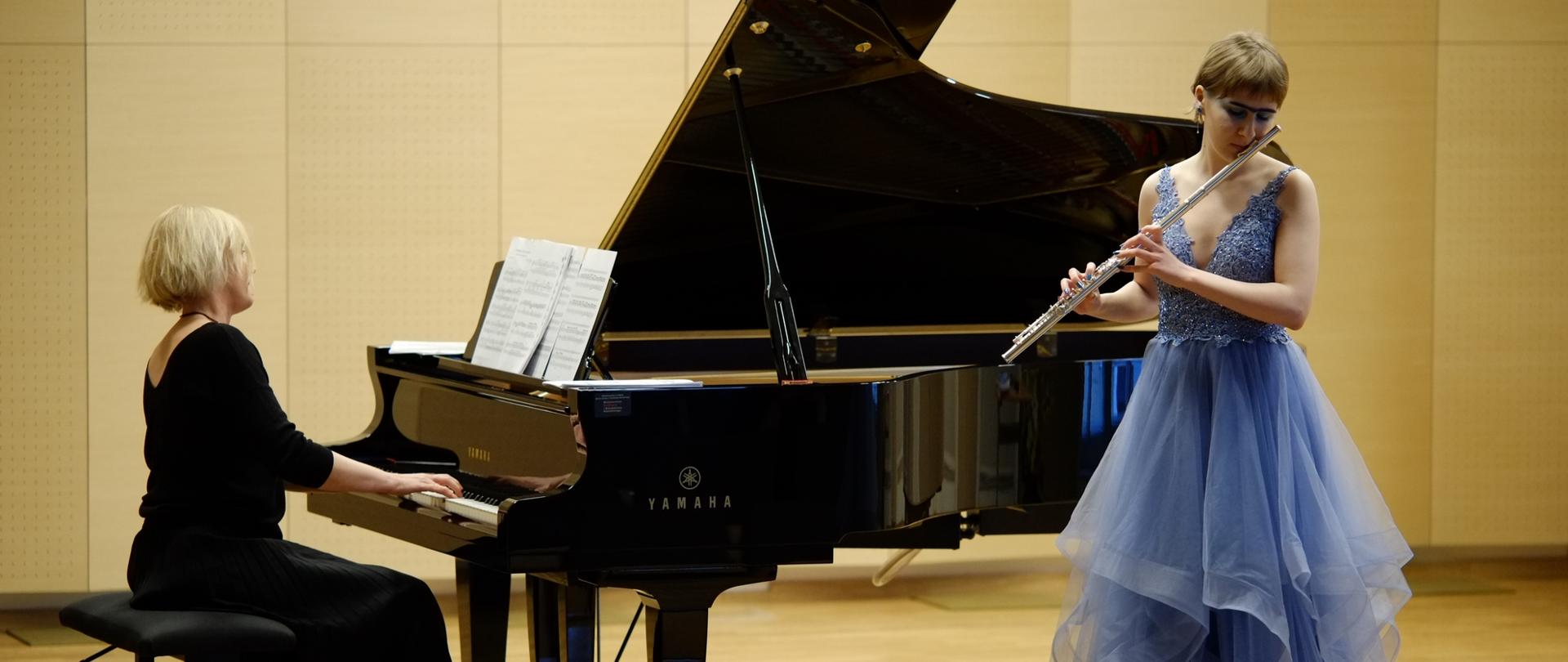 zdjęcie przedstawia uczennicę trzymającą przy twarzy flet poprzeczny oraz akompaniatora rozpoczynającego utwór na fortepianie na scenie sali koncertowej