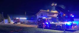 Strażacy gaszą pożar budynku jednorodzinnego z drabiny mechanicznej. W tle widać drugi pojazd pożarniczy biorący udział w akcji. 