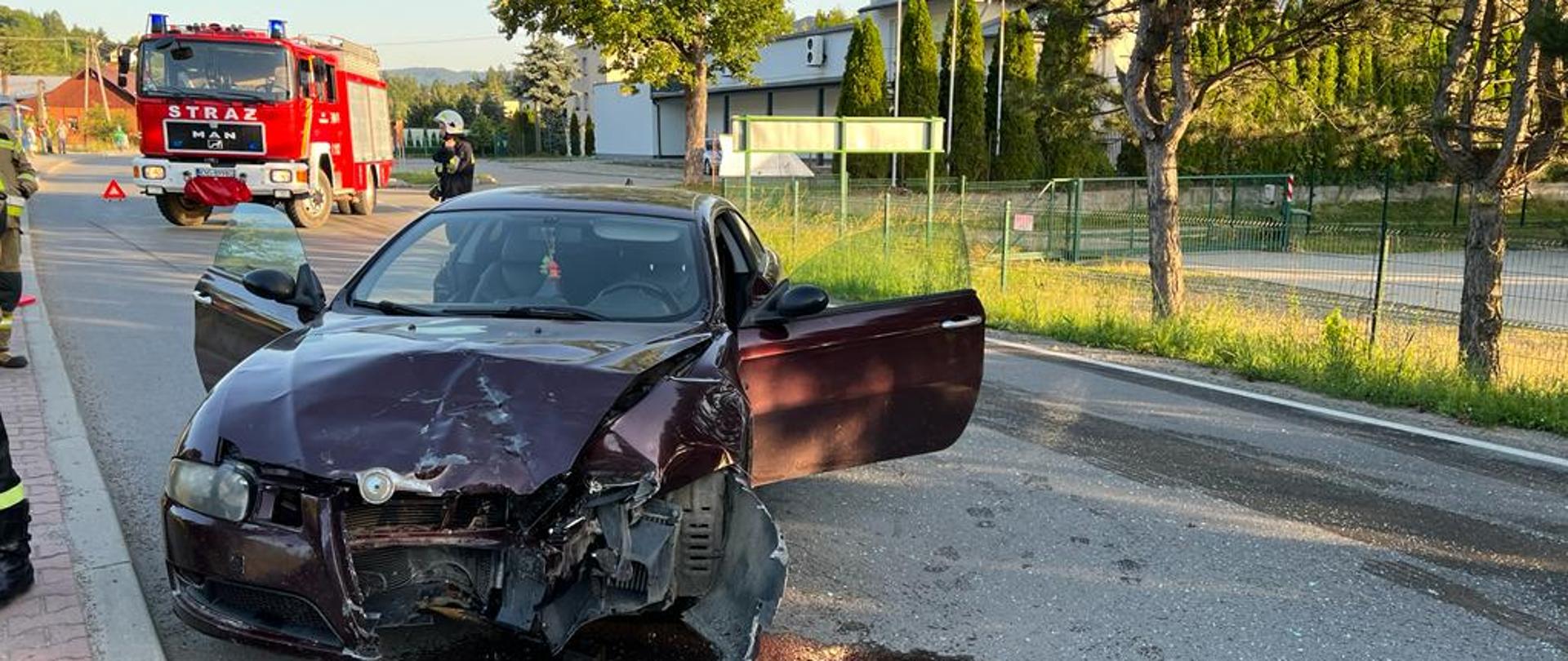 Rozbity samochód osobowy stoi pod skosem na drodze . W tle samochód pożarniczy.