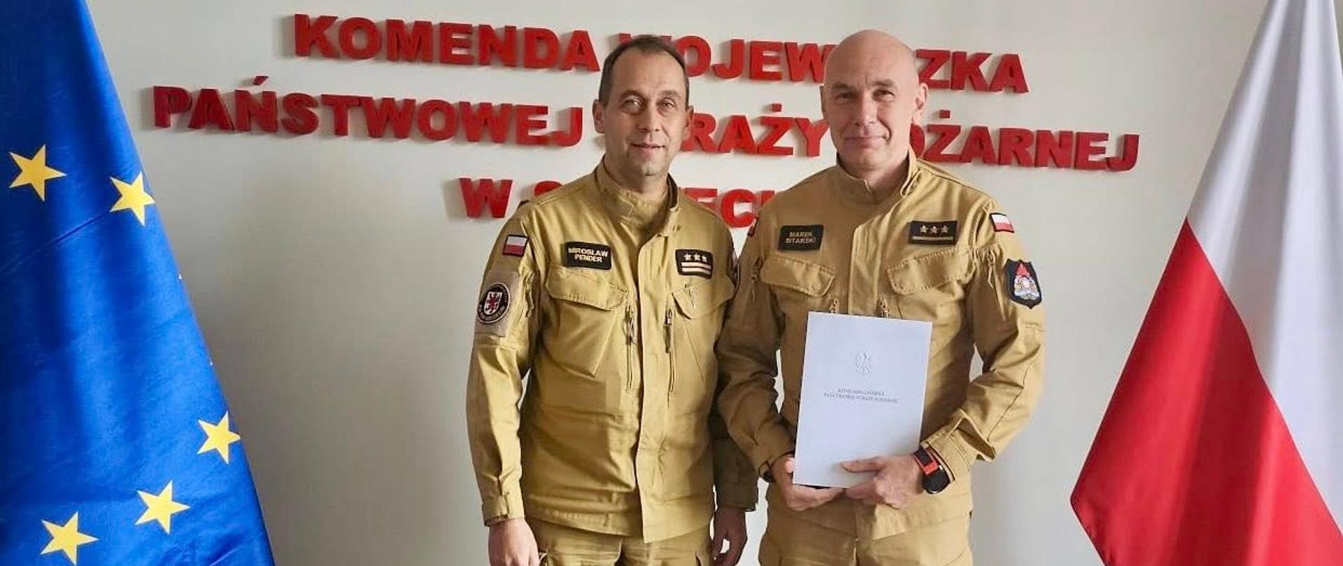 Na zdjęciu widać dwóch mężczyzn w mundurach koloru piaskowego, stojących obok siebie na tle ściany. Obaj mają krótkie włosy i patrzą prosto w obiektyw. Za nimi znajduje się ściana z czerwonym napisem i godłem Państwowej Straży Pożarnej (Państwowa Straż Pożarna). Po lewej stronie mężczyzn stoi flaga Unii Europejskiej, a po prawej flaga Polski.