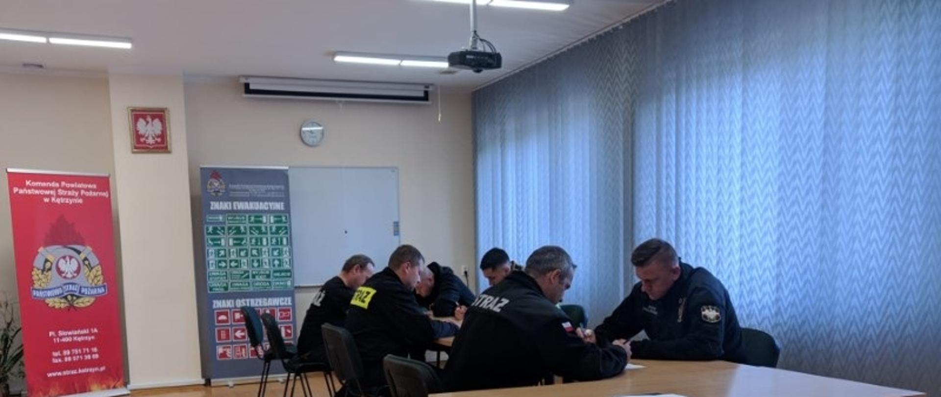 Sześciu strażaków siedzi przy stolikach i rozwiązuje test.