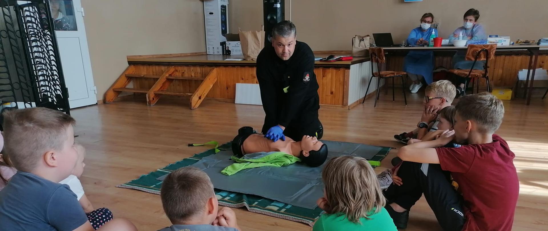 Zdjęcie przedstawia instruktaż pierwszej pomocy przy punkcie szczepień w Mącznikach. Na zdjęciu widzimy funkcjonariusza PSP, który przeprowadza pogadankę oraz ćwiczenia z pierwszej pomocy dla dzieci zgromadzonych na spotkaniu. Zdjęcie wykonane w porze dziennej w budynku sali OSP Mączniki.
