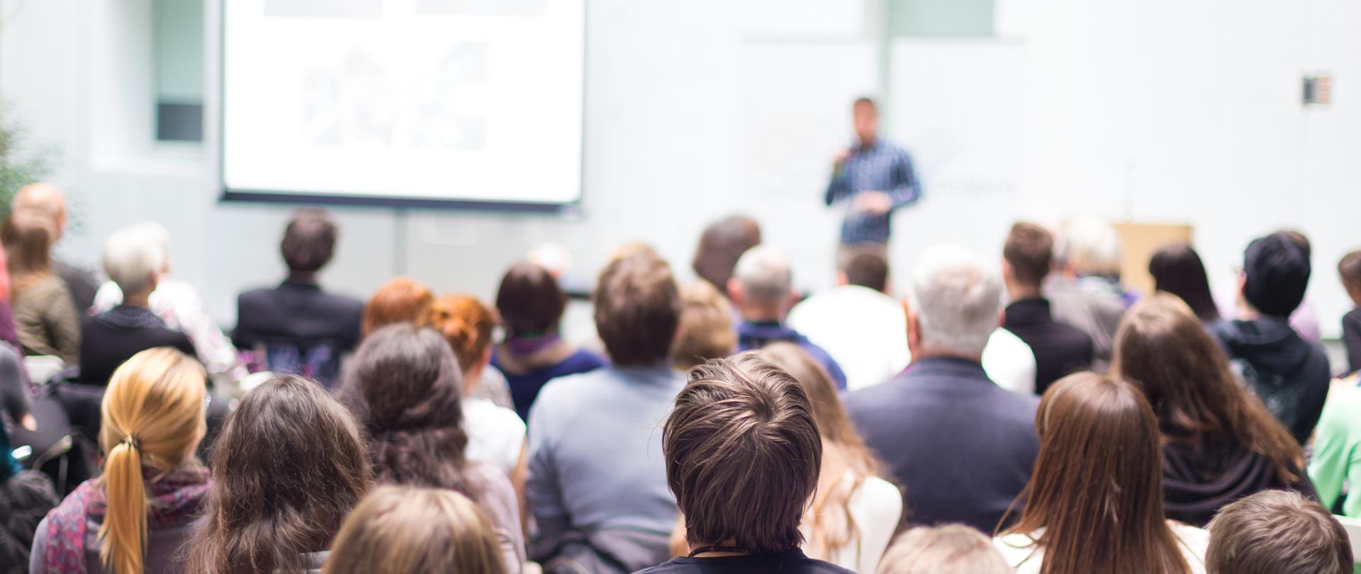 Wykładowca na sali konferencyjnej. Tyłem siedzi publiczność. Z lewej strony widoczna biała tablica.