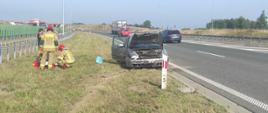Na pasie zieleni między jezdniami drogi S7 stoi rozbity samochód. Strażacy udzielają pomocy medycznej poszkodowanej.