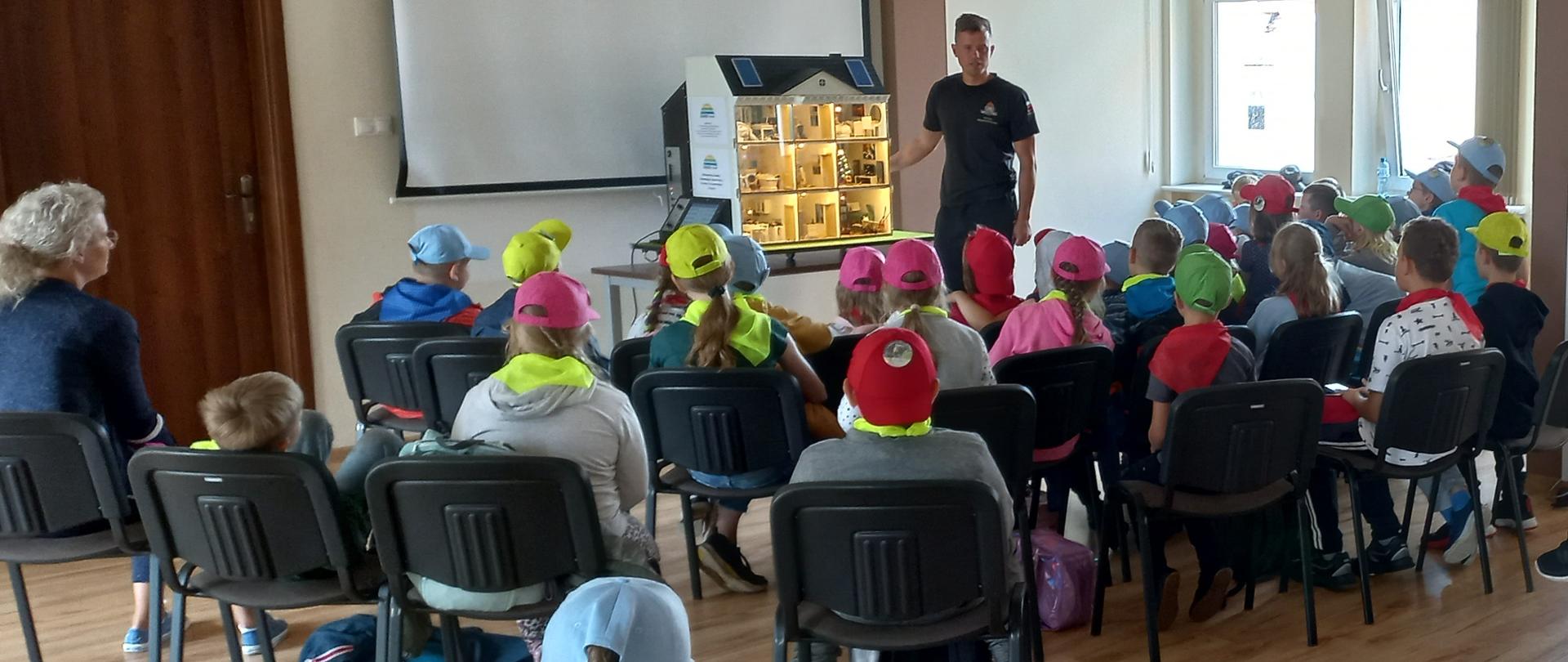 Zdjęcie przedstawia strażaków edukujących dzieci i młodzież, którzy wypoczywają w okresie wakacyjnym na koloniach i półkoloniach i zwiedzają siedzibę Jednostki Ratowniczo - Gaśniczej.
W tle świetlica Komendy.
