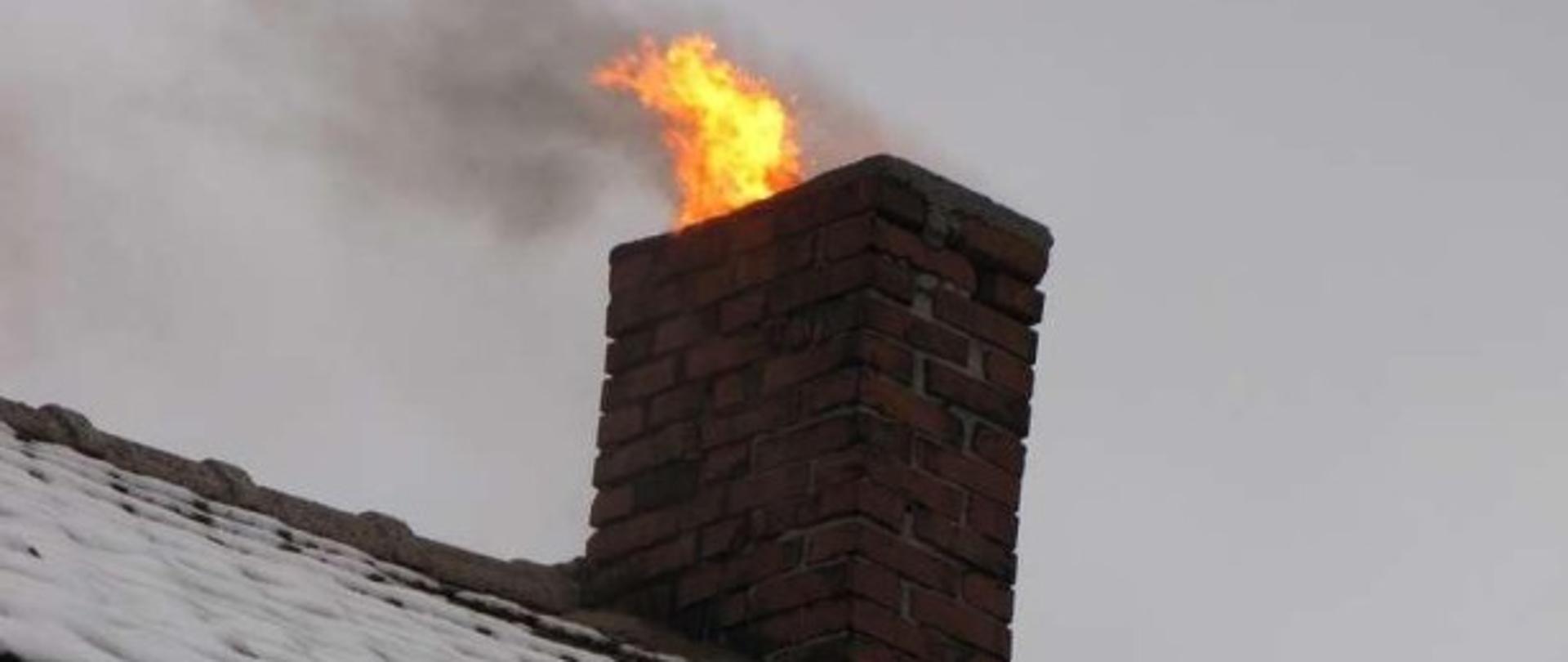 Na zdjęciu widać przysypaną warstwą śniegu połać dachu krytego dachówka ceramiczną, komin wykonany z cegły pełnej i wydobywający się z niego płomień ognia. 