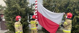 Na zdjęciu trzech strażaków mocuje flagę państwową na maszcie. ubrani są w ubrania specjalne bojowe koloru piaskowego, jasno żółty i czerwone hełmy.