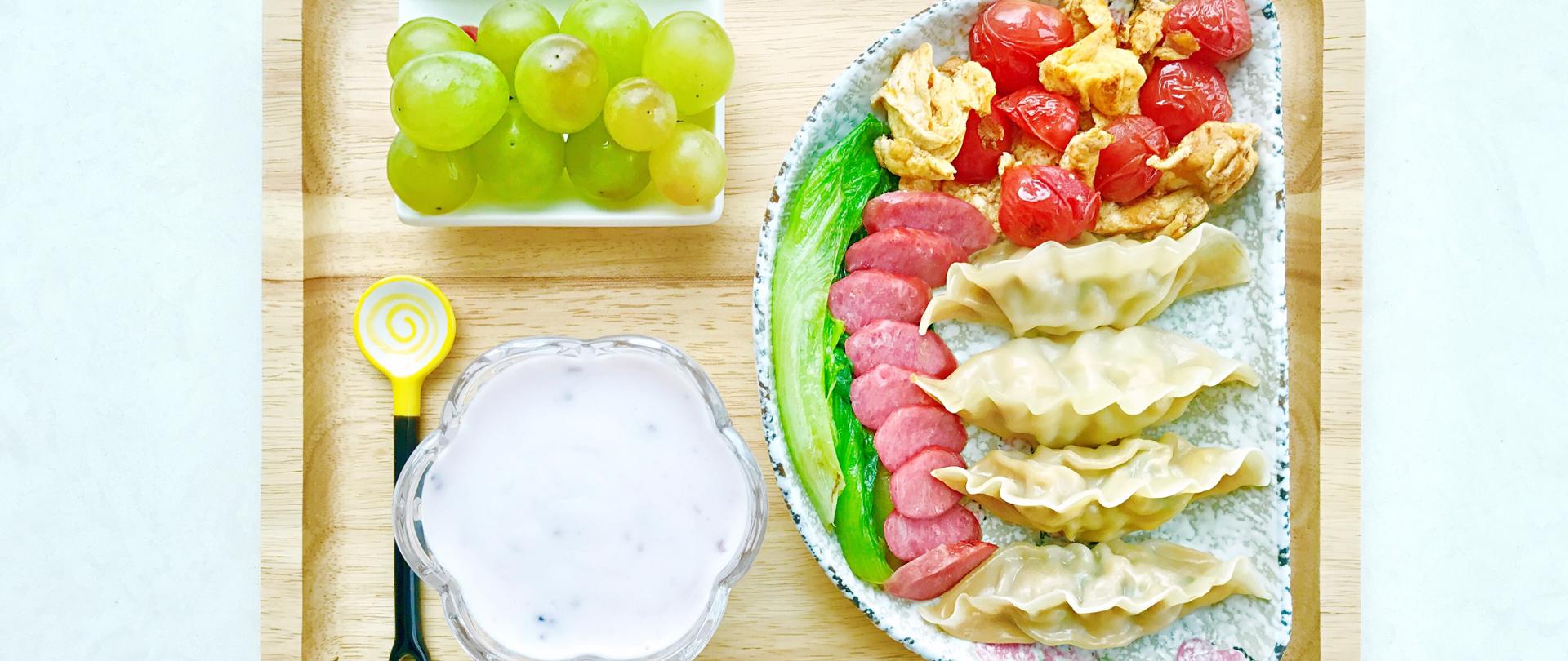 deska z jedzeniem, jogurt w misce, łyżka, winogrona, na talerzu pierogi, pomidory, pokrojona kiełbasa