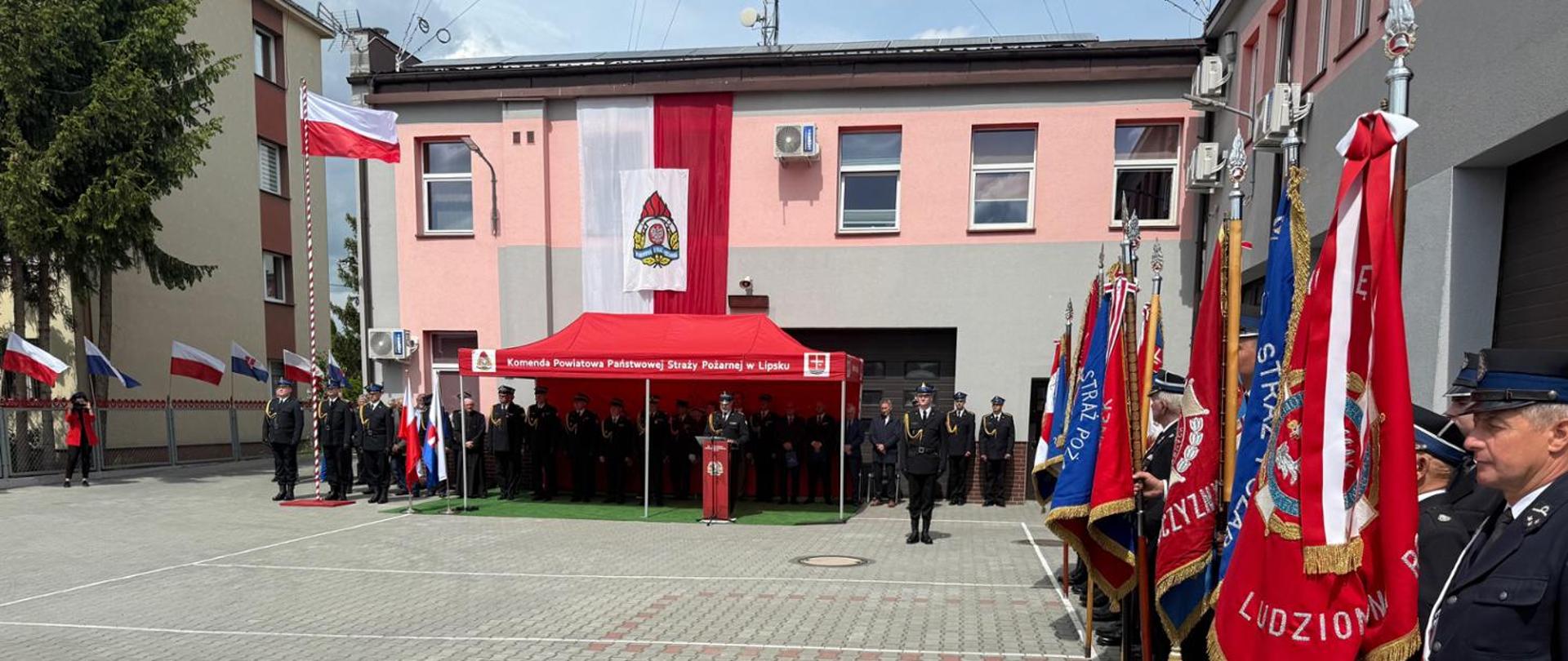 Widoka miejsca uroczystości strażackiej przed dużym budynkiem z rozwieszoną pionowo na ścianie flagą Polski i logotypem Państwowej Straży Pożarnej. Na placu przed budynkiem stoją poczty sztandarowe, czerwony namiot z mównicą, maszt z podniesioną flagą Polski. 
