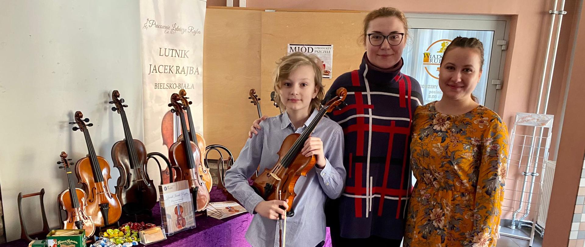 Zdjęcie przedstawiające stojącego ucznia - Antoniego Bulszę ze skrzypcami (po lewej stronie), akompaniatora - Liliię Tarasenko (na środku) i nauczyciela - panią Agatę Klich (po prawej stronie) w miejscu odbywania się III Ogólnopolskiego Konkursu „Co młodym w duszy gra?” w Rudzie Śląskiej.
