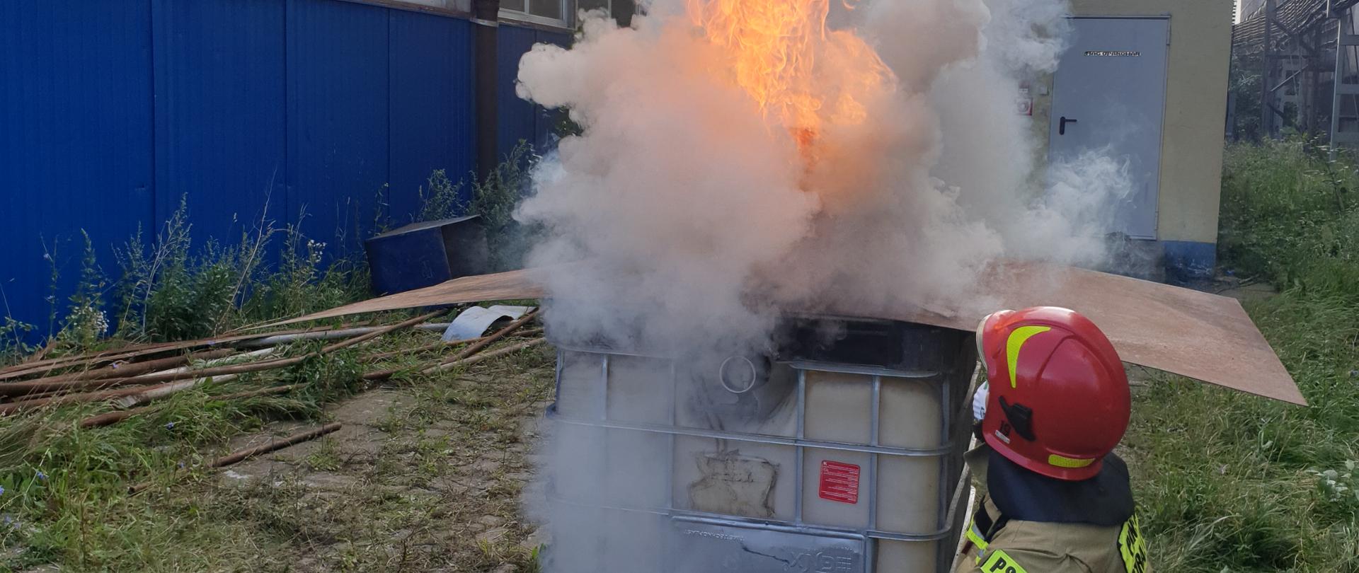 Strażak przedstawia zjawisko zapalenia gazów pożarowych na specjalnie do tego przygotowanym domku. 