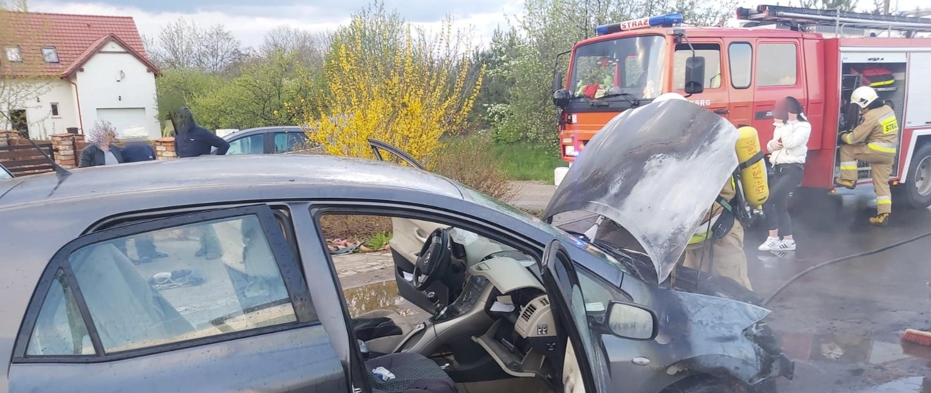 Na zdjęciu spalony samochód osobowy strażak w ODO oraz pojazd strażacki przy którym stoi ratownik