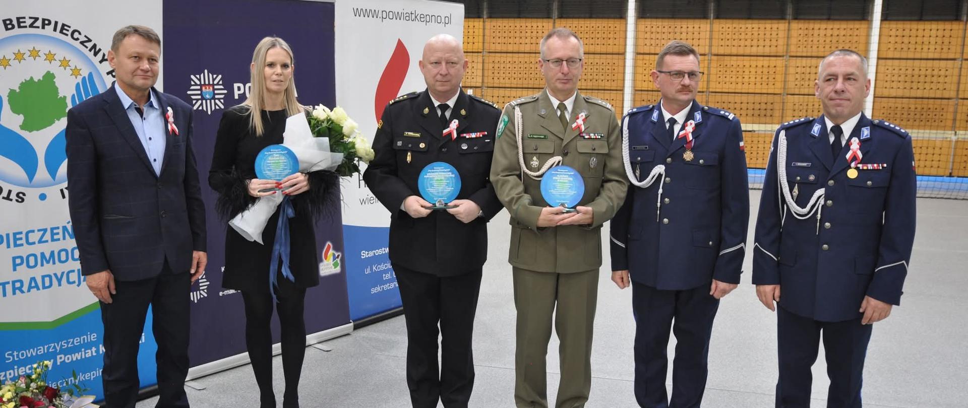 Grupa sześciu osób stojących na tle banerów Stowarzyszenia Nasz Bezpieczny Powiat Kępiński i Powiatu Kępińskiego. Wśród nich przedstawiciele służb mundurowych oraz samorządu, trzymający wyróżnienia i kwiaty.