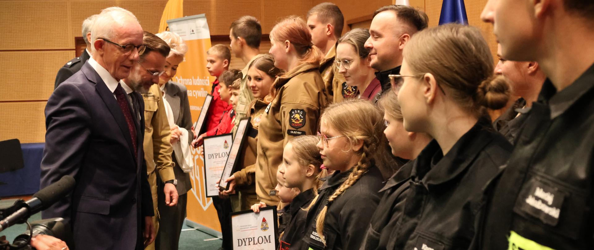 Uroczyste wręczenie nagród laureatom konkursu „Budujemy SYSTEM ODPORNOŚCI SPOŁECZNEJ” - wyróżnieni laureaci stoją na podeście sali w Podlaskim Urzędzie Wojewódzkim, organizatorzy wydarzenia wręczają dyplomy i gratulują