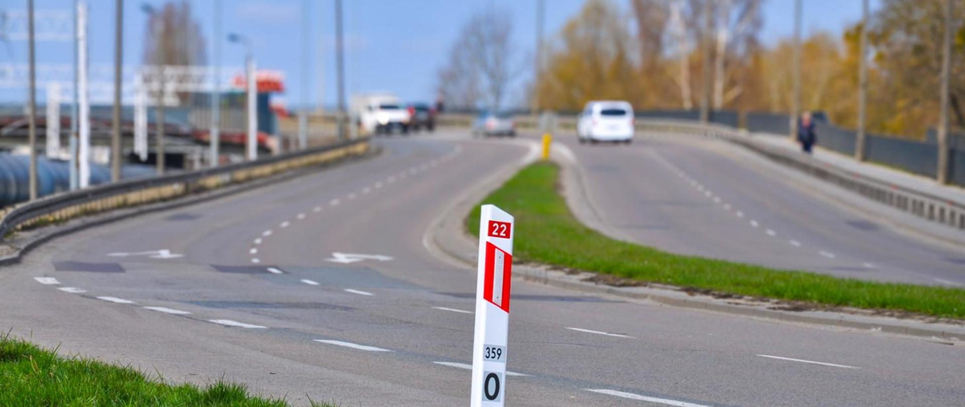 Dwupasmowa droga krajowa z rozdzielającym pasem zieleni; na pierwszym planie biało‑czerwony słupek kilometrowy z oznaczeniem „22” i „359”, w tle kilka samochodów jadących w obu kierunkach, barierki drogowe oraz drzewa bez liści przy jezdni.