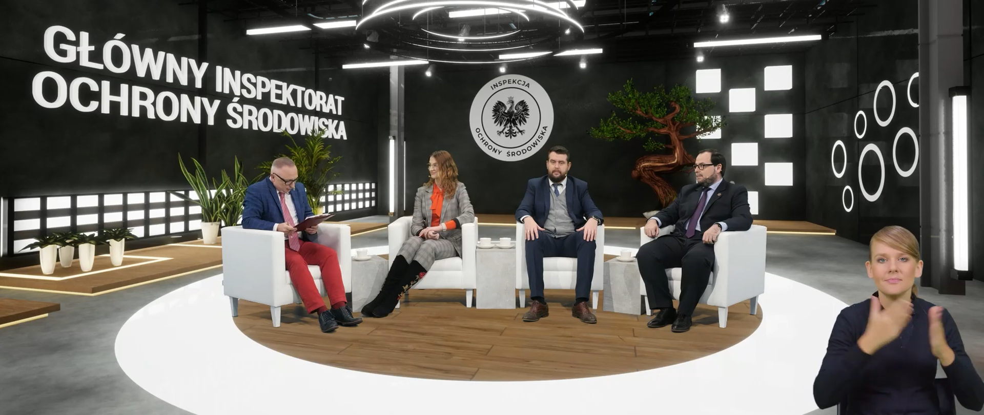 Trzech mężczyzn oraz kobieta siedzą na białych fotelach. Jeden z mężczyzn czyta coś z kartki, pozostali patrzą się na niego. Tło jest w czarno białej tonacji. Znajduje się na nim napis i logo GIOŚ