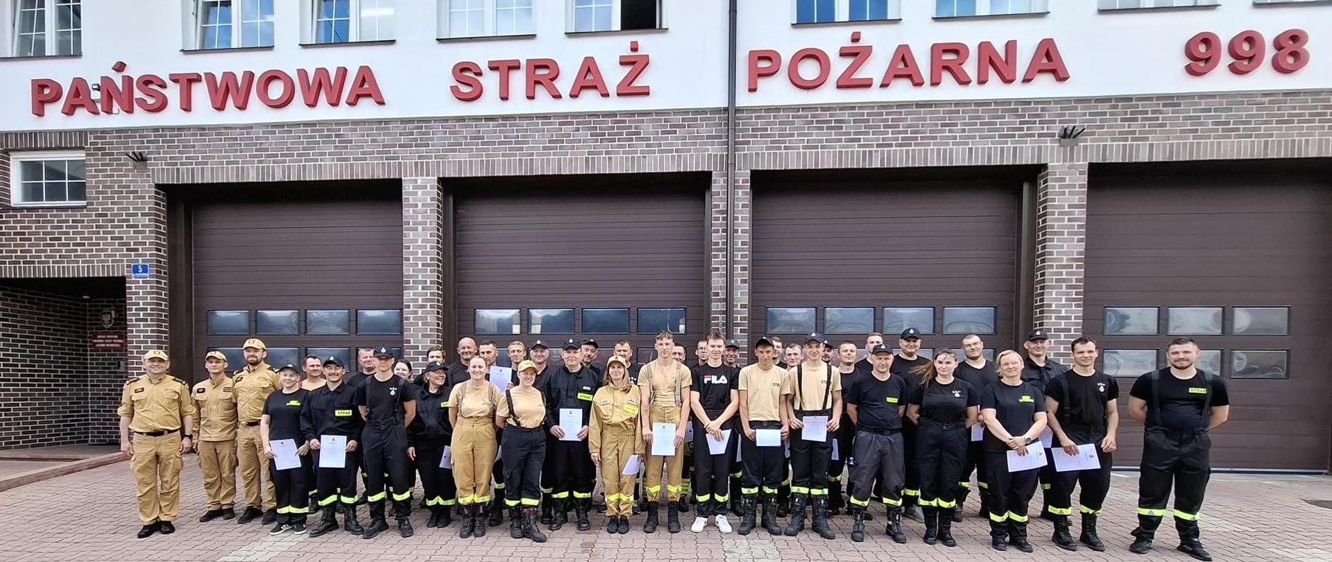 Na zdjęciu widać dużą grupę około 40 osób, młodych dorosłych i dorosłych, ustawionych w rzędach przed budynkiem straży pożarnej. Większość z nich ubrana jest w mundury strażackie w różnych kolorach – jasnobeżowym (piaskowym), granatowym lub czarnym. Niektórzy trzymają w rękach białe kartki, certyfikaty ukończenia szkolenia. Budynek za nimi ma ceglane ściany na parterze i jaśniejszą elewację na piętrze, z widocznymi dużymi, przeszklonymi oknami. Na parterze znajdują się trzy duże, brązowe bramy garażowe. Nad bramami widnieje napis "PAŃSTWOWA STRAŻ POŻARNA", a po prawej stronie numer "998". W tle widać fragmenty nieba, które jest częściowo zachmurzone.