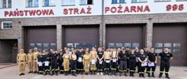Na zdjęciu widać dużą grupę około 40 osób, młodych dorosłych i dorosłych, ustawionych w rzędach przed budynkiem straży pożarnej. Większość z nich ubrana jest w mundury strażackie w różnych kolorach – jasnobeżowym (piaskowym), granatowym lub czarnym. Niektórzy trzymają w rękach białe kartki, certyfikaty ukończenia szkolenia. Budynek za nimi ma ceglane ściany na parterze i jaśniejszą elewację na piętrze, z widocznymi dużymi, przeszklonymi oknami. Na parterze znajdują się trzy duże, brązowe bramy garażowe. Nad bramami widnieje napis "PAŃSTWOWA STRAŻ POŻARNA", a po prawej stronie numer "998". W tle widać fragmenty nieba, które jest częściowo zachmurzone.