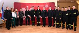 awansowani strażacy na tle ścianki w obecności innych funkcjonariuszy i pracowników cywilnych