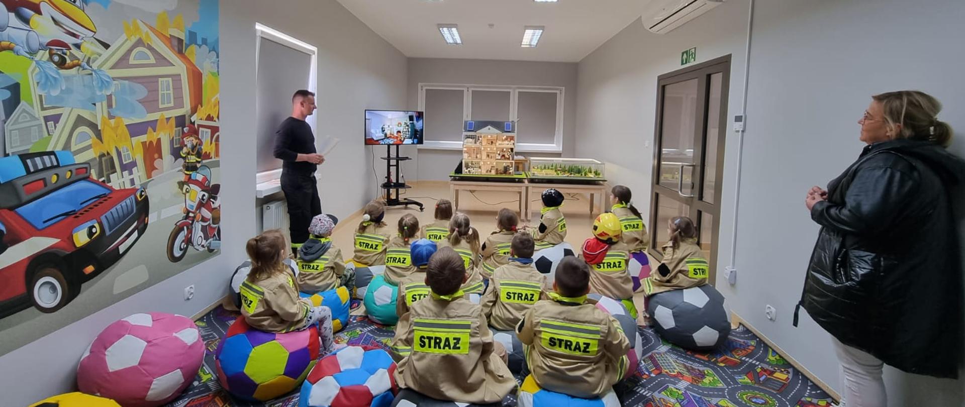 Dzieci siedzą w salce edukacyjnej. Strażak opowiada.