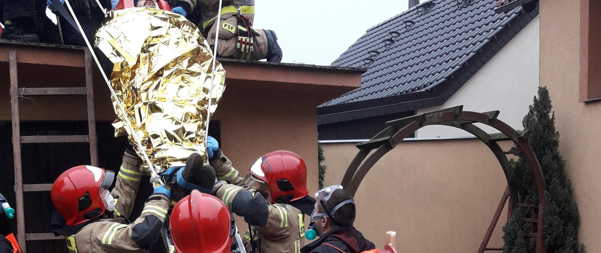 Ewakuacja poszkodowanego z dachu przy użyciu opartej o budynek drabiny. Po drabinie strażacy wraz z ratownikami medycznymi zsuwają nosze, zabezpieczając je liną.
