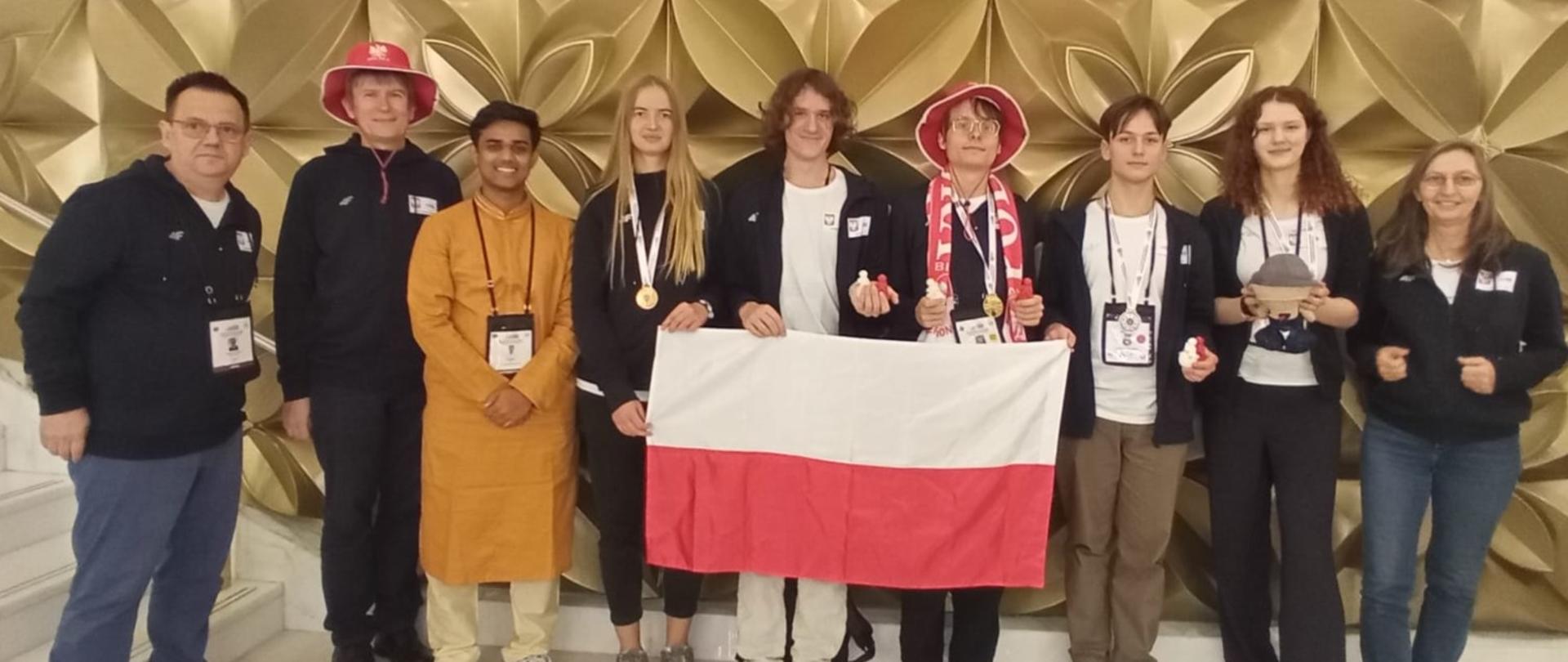 Grupa dziewięciu osób, w tym młodzież i dorośli, stoi w rzędzie przed złotą, wzorzystą ścianą. Cztery osoby trzymają polską flagę. Młoda kobieta w czarnej bluzie i dżinsach, po prawej stronie grupy, trzyma pluszaka. Dwie osoby mają na głowie biało-czerwone kapelusze. Wszyscy z wyjątkiem jednej osoby mają na szyi medale na wstążkach. Po lewej i prawej stronie grupy stoją dorośli w ciemnych strojach z logami. Całość wygląda na oficjalne zdjęcie z zawodów lub olimpiady.