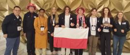 Grupa dziewięciu osób, w tym młodzież i dorośli, stoi w rzędzie przed złotą, wzorzystą ścianą. Cztery osoby trzymają polską flagę. Młoda kobieta w czarnej bluzie i dżinsach, po prawej stronie grupy, trzyma pluszaka. Dwie osoby mają na głowie biało-czerwone kapelusze. Wszyscy z wyjątkiem jednej osoby mają na szyi medale na wstążkach. Po lewej i prawej stronie grupy stoją dorośli w ciemnych strojach z logami. Całość wygląda na oficjalne zdjęcie z zawodów lub olimpiady.