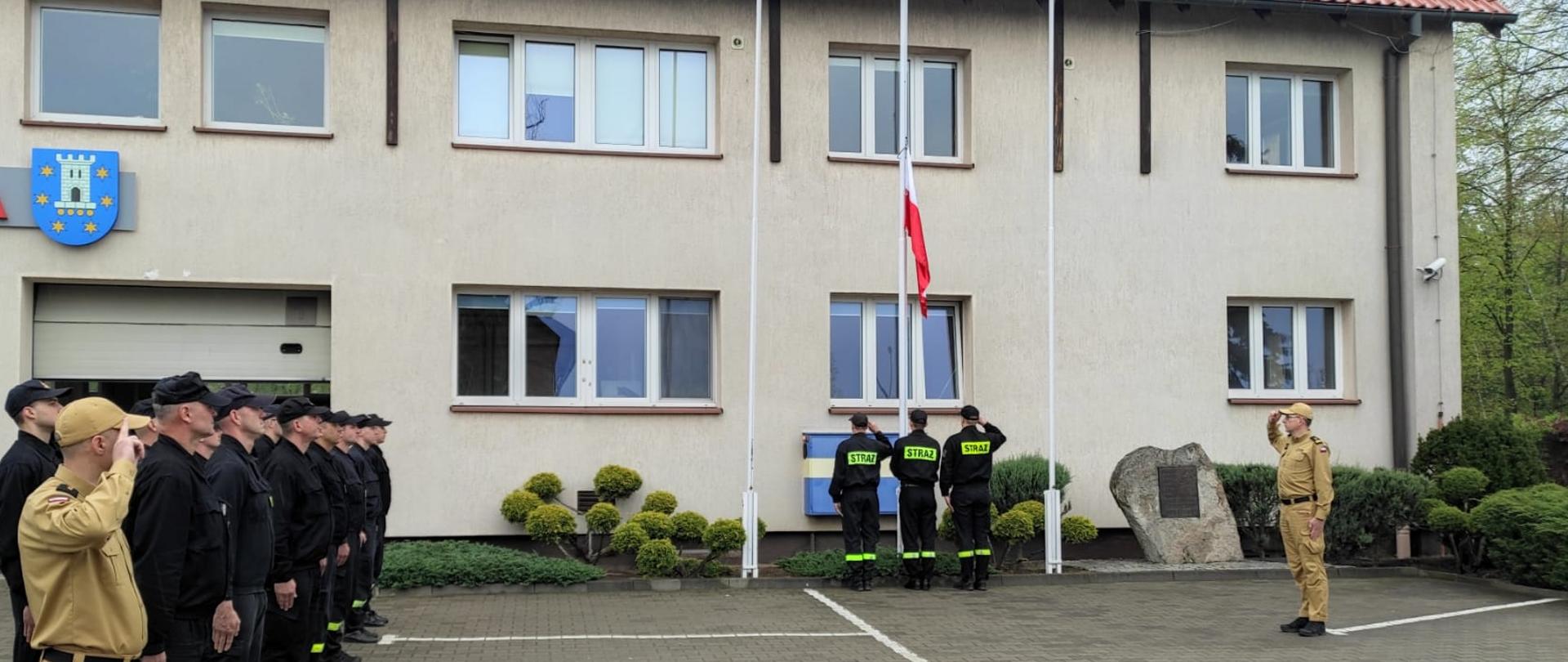 Pleszewscy strażacy PSP podczas uroczystego podniesienia plagi państwowej. Zdjęcę zrobione przed budynkiem komendy PSP.. Po lewej stronie widoczni strażacy ustawieni w dwuszeregu, w centrum widoczny maszt i poczet flagowy dokonujący uroczystego podniesienia, po prawej stronie salutujący dowódca uroczystości