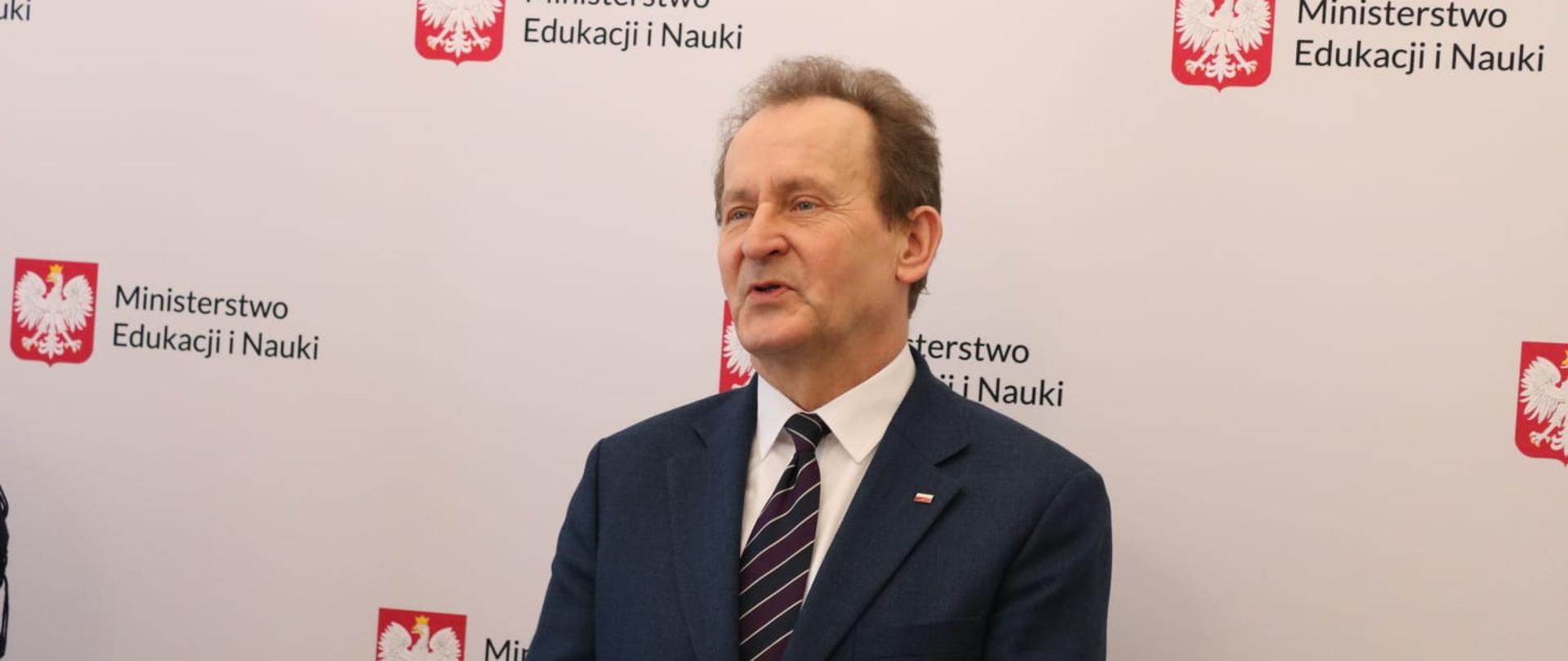 Wiceminister Włodzimierz Bernacki podczas uroczystości, za nim stoi baner z napisem Ministerstwo Edukacji i Nauki.