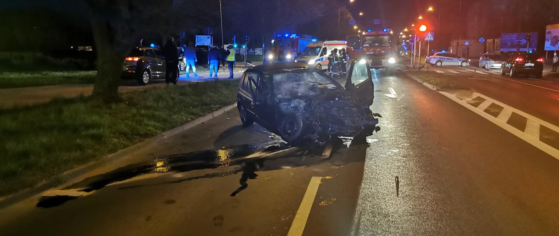Zdjęcie przedstawia dwa samochody osobowe po zderzeniu. Jeden z pojazdów stoi na jezdni z uszkodzonym przodem. Drugi pojazd stoi na chodniku . Na zdjęciu widać samochody strażackie, radiowozy oraz karetkę pogotowia ratunkowego. Ponadto na zdjęciu widać policjantów i strażaków. Pora nocna. 