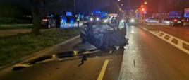 Zdjęcie przedstawia dwa samochody osobowe po zderzeniu. Jeden z pojazdów stoi na jezdni z uszkodzonym przodem. Drugi pojazd stoi na chodniku . Na zdjęciu widać samochody strażackie, radiowozy oraz karetkę pogotowia ratunkowego. Ponadto na zdjęciu widać policjantów i strażaków. Pora nocna. 