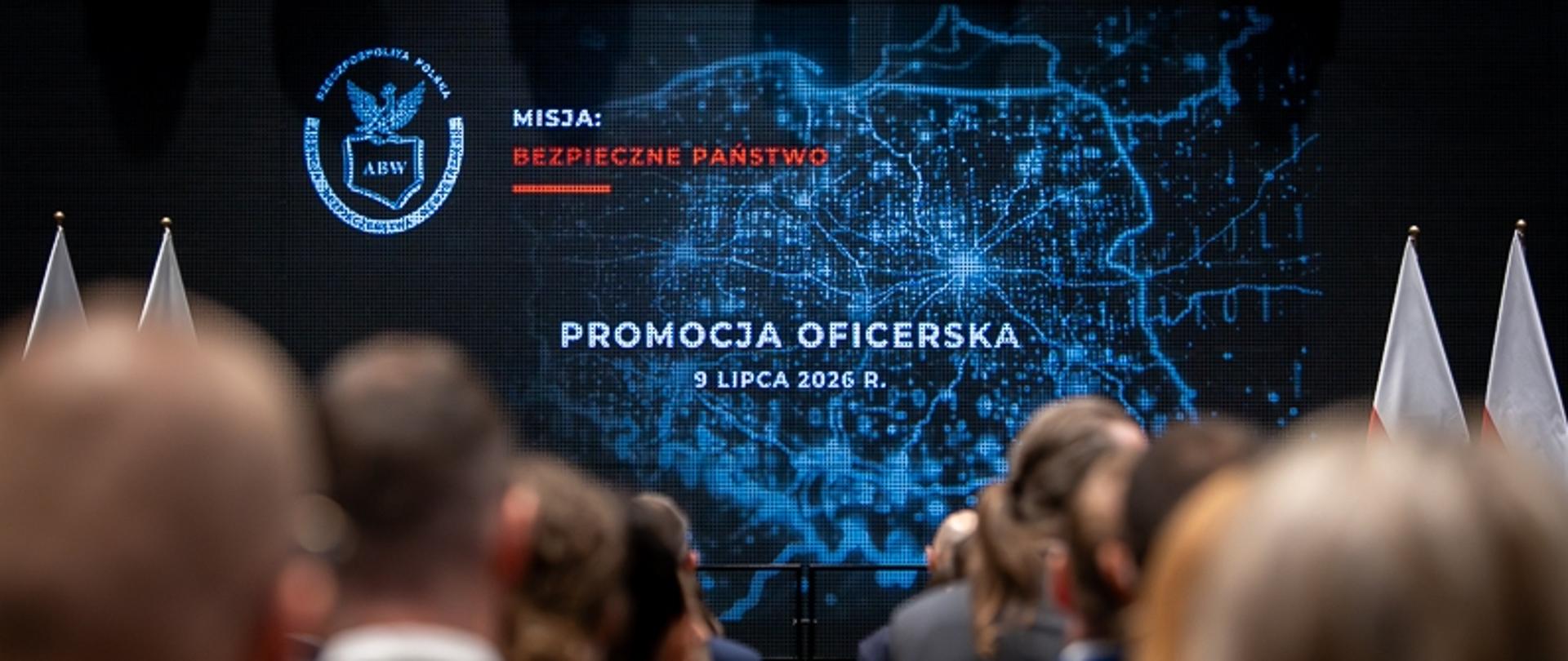 Zdjęcie przedstawia promocję funkcjonariuszek i funkcjonariuszy Agencji Bezpieczeństwa Wewnętrznego na pierwszy stopień oficerski. Promowani stoją tyłem. W tle na ekranie wyświetlone kontury Polski z podświetlonymi miastami i drogami oraz napisem na środku: PROMOCJA OFICERSKA 9 LIPCA 2026 R. Po lewej stronie na górze wyświetlono logo ABW (orzeł trzymający kontur Polski z napisem ABW) oraz napis: MISJA: BEZPIECZNE PAŃSTWO. U góry zdjęcia widać również czarne reflektory. Po prawej i lewej stronie ekranu widać stojące biało-czerwone flagi Polski - po dwie z każdej strony.