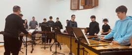 Koncert klasy perkusji - zdjęcie przedstawia grupę młodych muzyków grających na różnych instrumentach na pierwszym planie marimby, za pulpitem nauczyciel prowadzący zespół. 