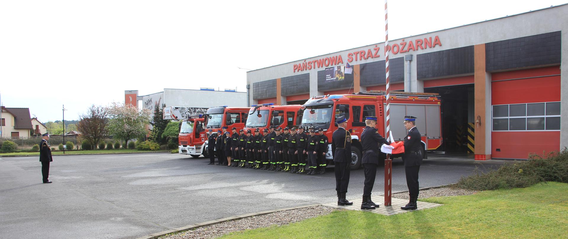 Trzech strażaków zawiesza Flagę Państwową na maszcie. Obok w szeregu stoją strażacy oraz samochody strażackie.