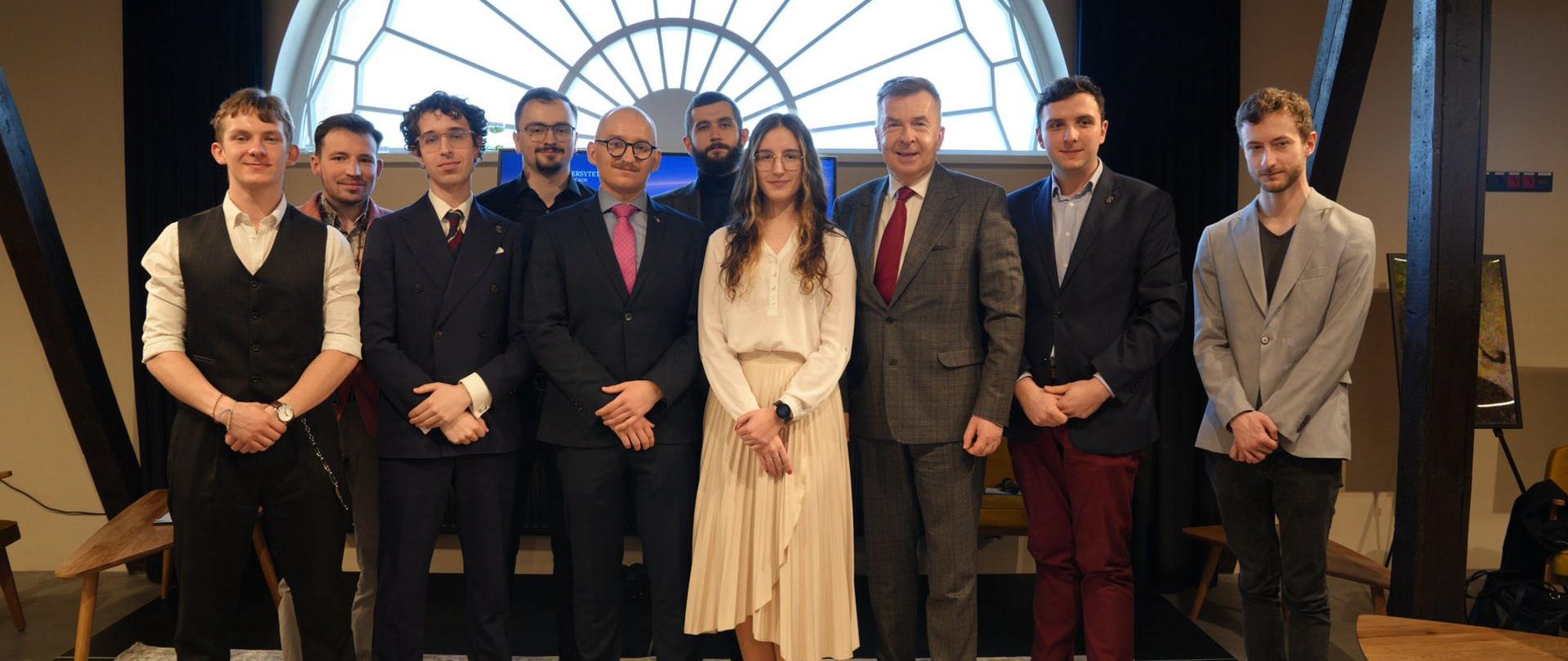 Na tle dużego półokrągłego okna stoi grupa dziesięciu osób, pośrodku minister Wieczorek.