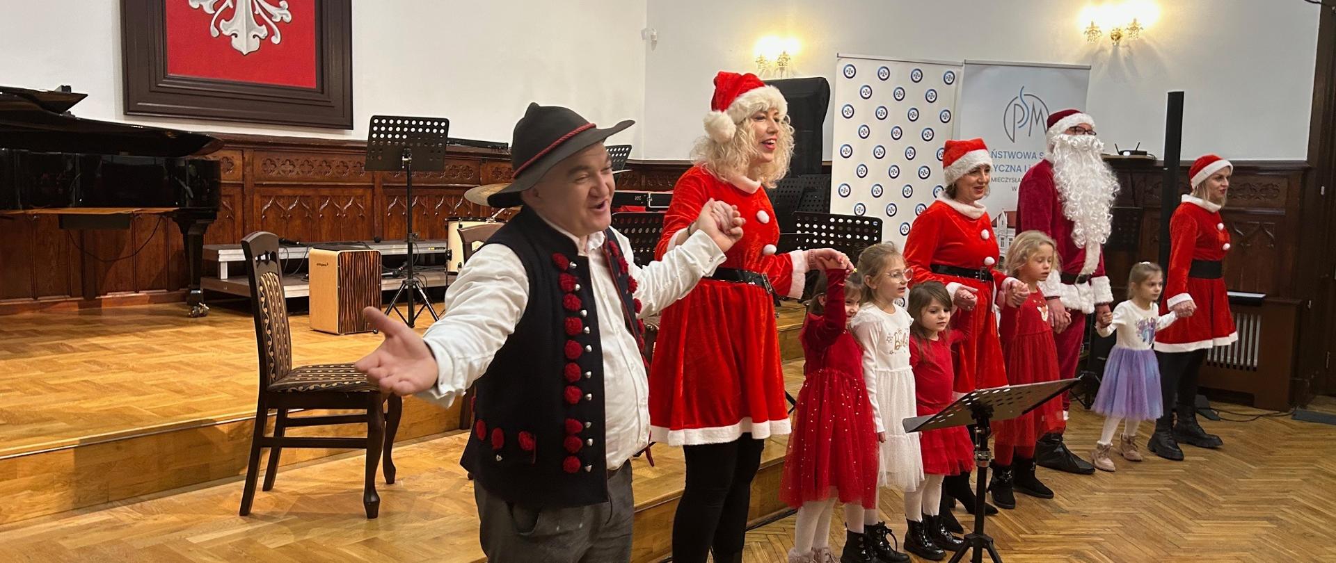 Uczniowie i nauczyciele wraz z dziećmi występują na scenie podczas finałowego koncertu charytatywnego, w świątecznych strojach, w sali koncertowej szkoły.