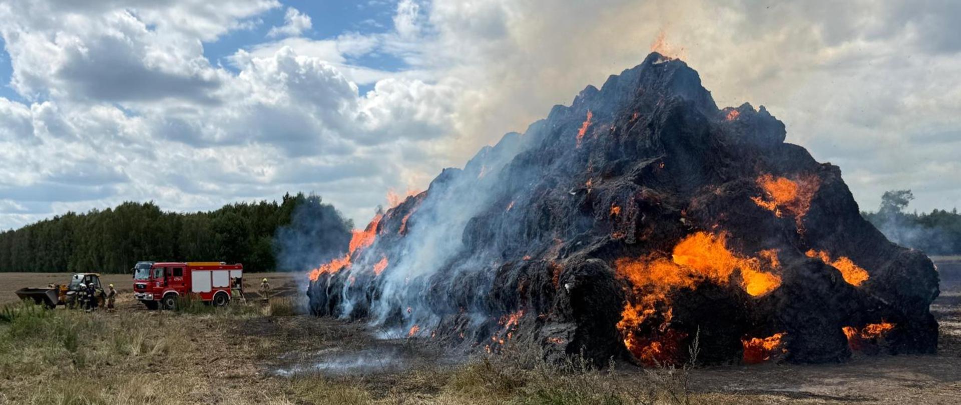 Wysoka hałda palących się belotów słomy, w tle samochód pożarniczy i strażacy gaszą pożar.