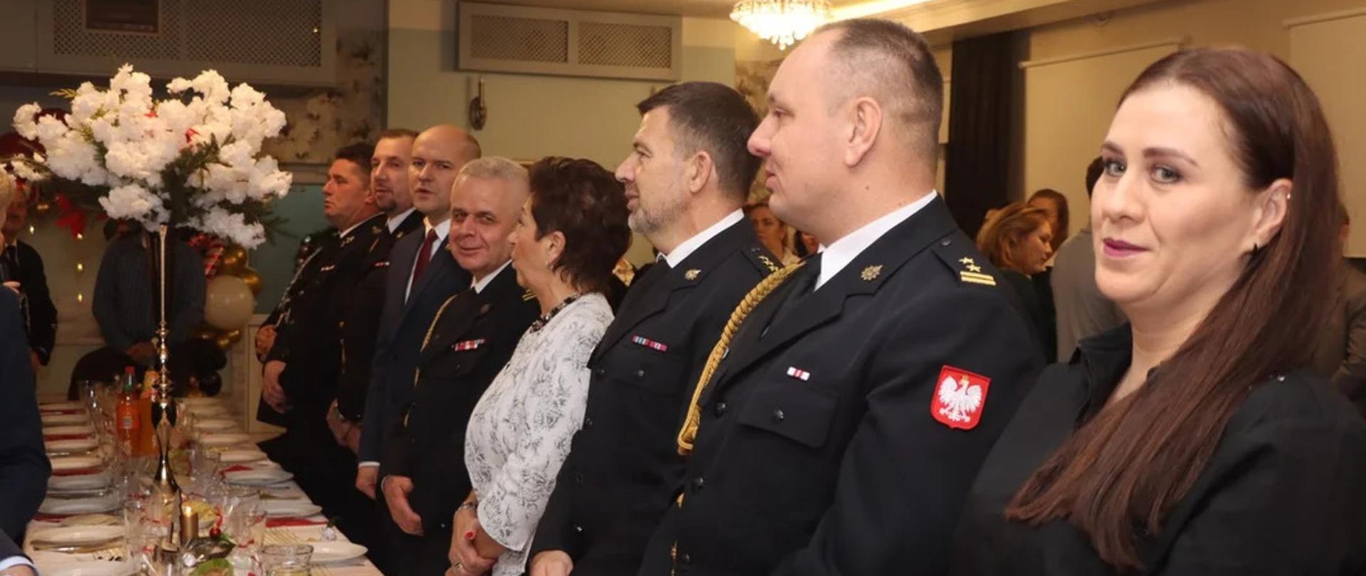 Wnętrze sali wiejskiej. Po obu stronach stołu, na którym znajduje się zastawa, napoje oraz potrawy, a także wazon z bukietem białych kwiatów, stoja uczestnicy spotkania. Są wsród nich strażacy PSP i OSP w mundurach wyjściowych oraz osoby cywilne (kobiety i mężczyźni)