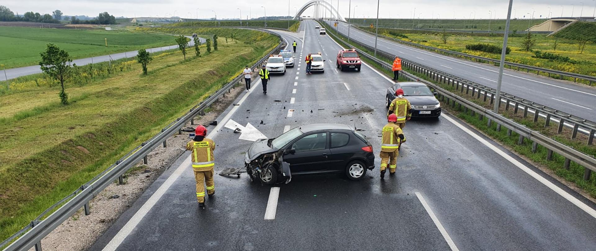 Na zdjęciu widać rozbity pojazd na drodze trzypasmowej. Przy samochodach znajdują się trzech strażaków w ubraniu koloru piaskowego. W tle widać również inne samochody