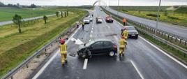 Na zdjęciu widać rozbity pojazd na drodze trzypasmowej. Przy samochodach znajdują się trzech strażaków w ubraniu koloru piaskowego. W tle widać również inne samochody