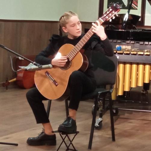 Scena z podłogą koloru brązowego, na środku uczennica ubrana na czarno siedzi ja krześle, gra na gitarze. Przed nią mikrofon na statywie, za nią fragment marimby. Ściany koloru białego i brązowego. Z tyłu wisi portret Paderewskiego. 