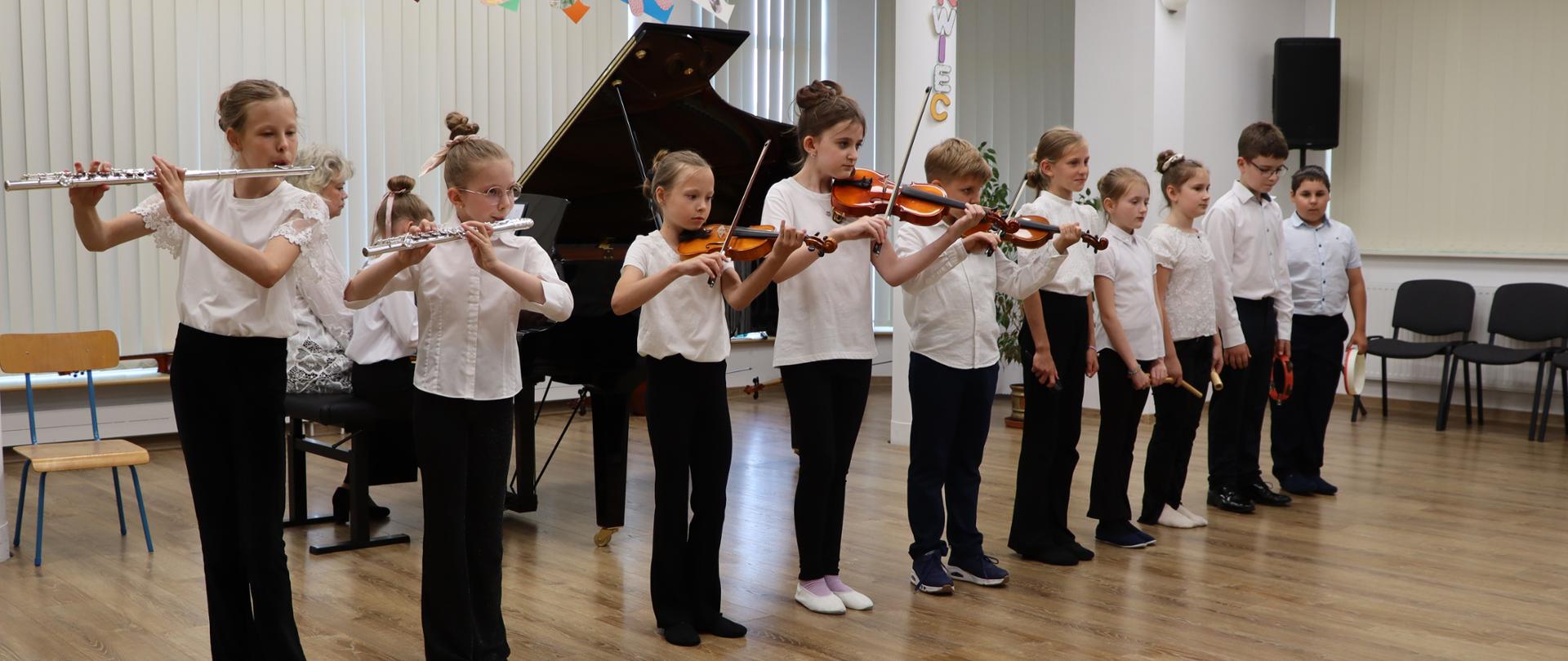 Koncert. Na środku sali koncertowej stoją uczniowie szkoły muzycznej. Dzieci ubrane są w białe koszule i czarne spodnie. Niektóre z nich grają na skrzypcach i fletach poprzecznych. 