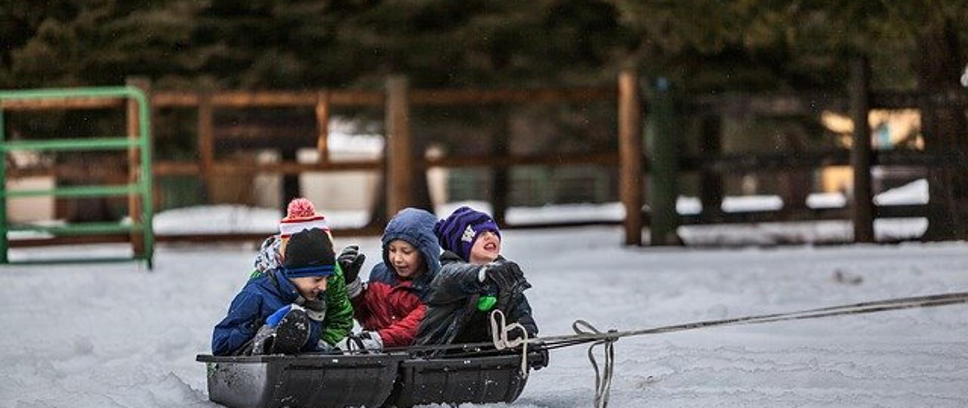 Zdjęcie przedstawia czterech uśmiechniętych chłopców, którzy znajdują na zewnątrz w zimowy dzień. Każdy z nich ubrany jest w ciepły strój. Wszyscy razem siedzą na czarnych sankach i dobrze się bawią na świeżym powietrzu. W tle znajdują się drewniane ogrodzenie, a za nim drzewa. 