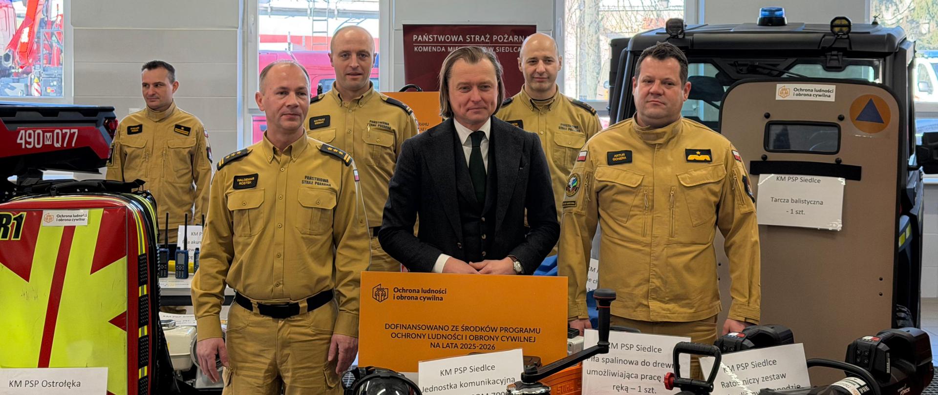 Do zdjęcia pozują uczestnicy wydarzenia - strażacy PSP w piaskowych mundurach służbowych z Mazowieckiem Komendantem Wojewódzkim PSP nadbryg. Arturem Gonerą i Komendantem Miejskim PSP w Siedlcach st. bryg. Waldemarem Rostkiem oraz Wojewodą Mazowieckim Mariuszem Frankowskim w garniturze na czele.