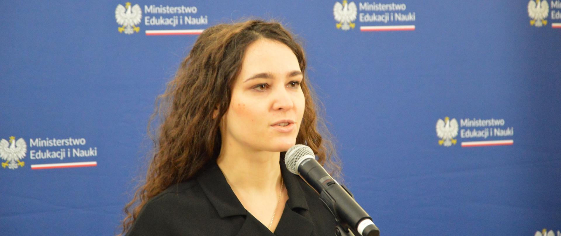 Młoda kobieta z długimi włosami ubrana na czarno stoi i mówi do mikrofonu na stojaku, za nią niebieska ściana z napisem Ministerstwo Edukacji i Nauki.