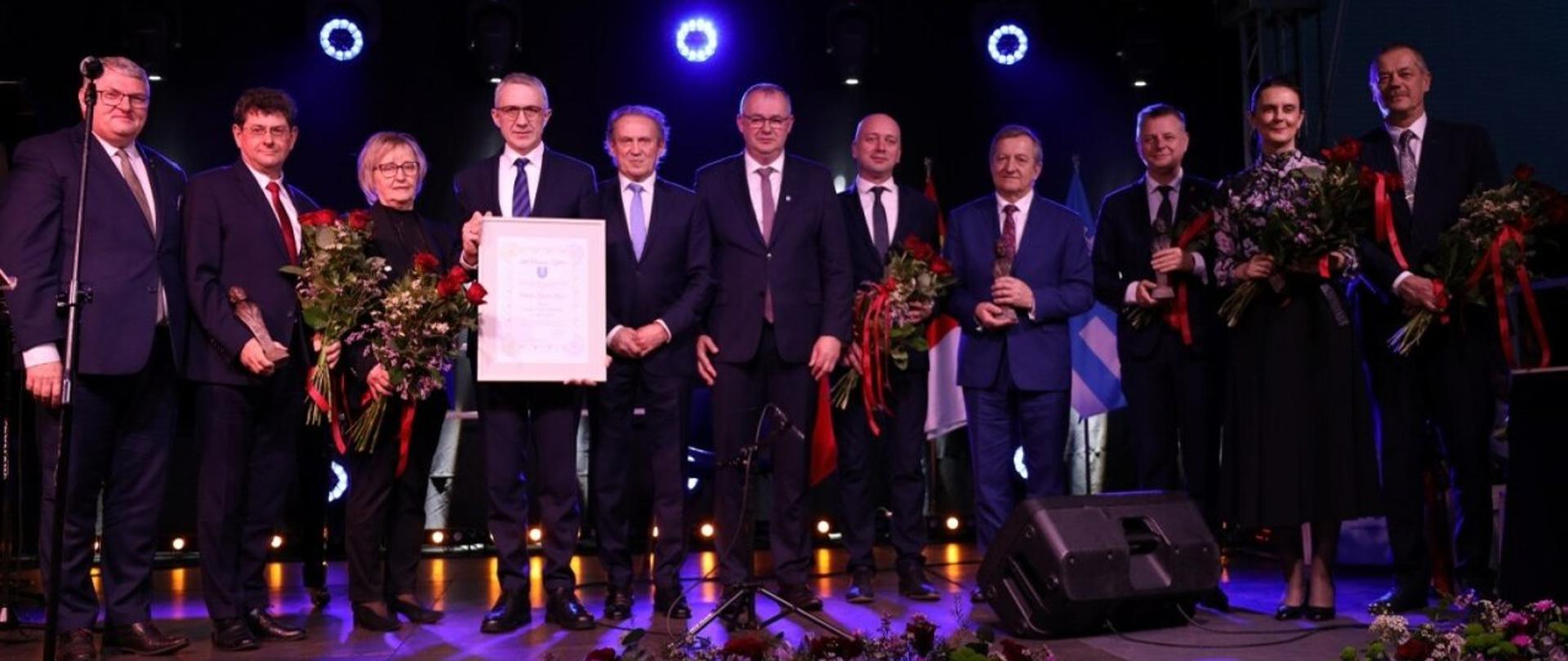 Na scenie stoi grupa meżczyzn i dwie kobiety, część z nich trzyma kwiaty, wszyscy ubrani są w stroje wyjściowe, jeden z mężczyzn trzyma w ręku duży dokument w ramie