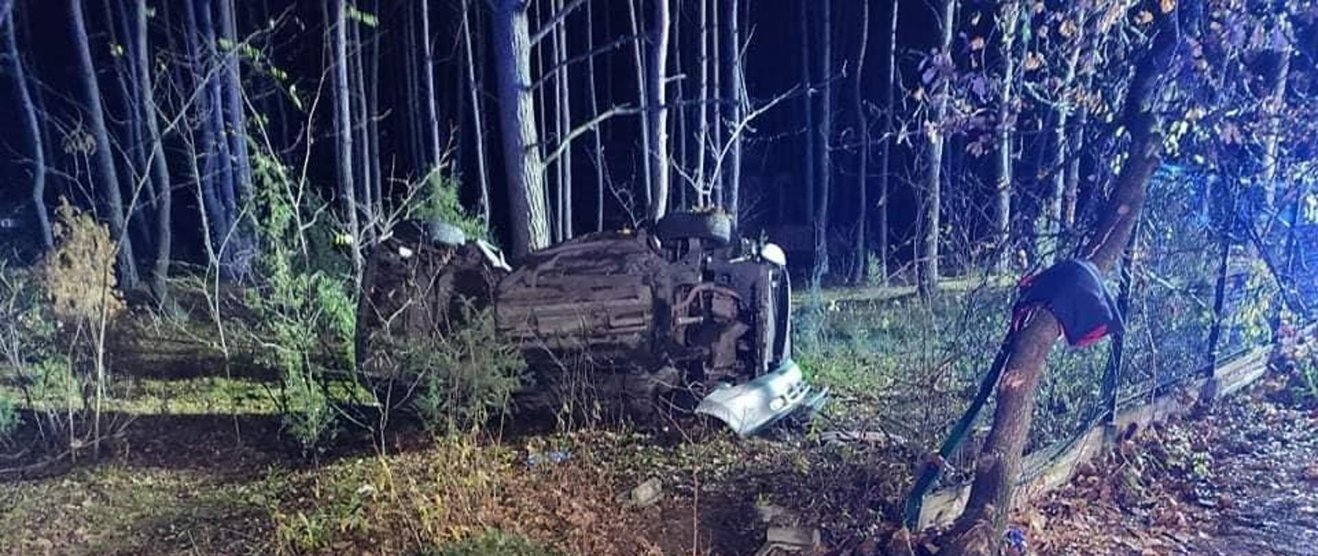 Na środku zdjęcia w oddali widoczny jest spód leżącego na boku samochodu osobowego. W środkowej części widoczne jest zniekształcenie powstałe w skutek uderzenia pojazdu w drzewo. Samochód znajduje się na skraju lasu, przed samochodem widoczne są leżące elementy pojazdu oraz uszkodzonego ogrodzenia z betonowa podmurówką, które uległy zniszczeniu w wyniku zdarzenia. W oddali widoczne drzewa, zaś w prawym dolnym rogu skrawek drogi asfaltowej.